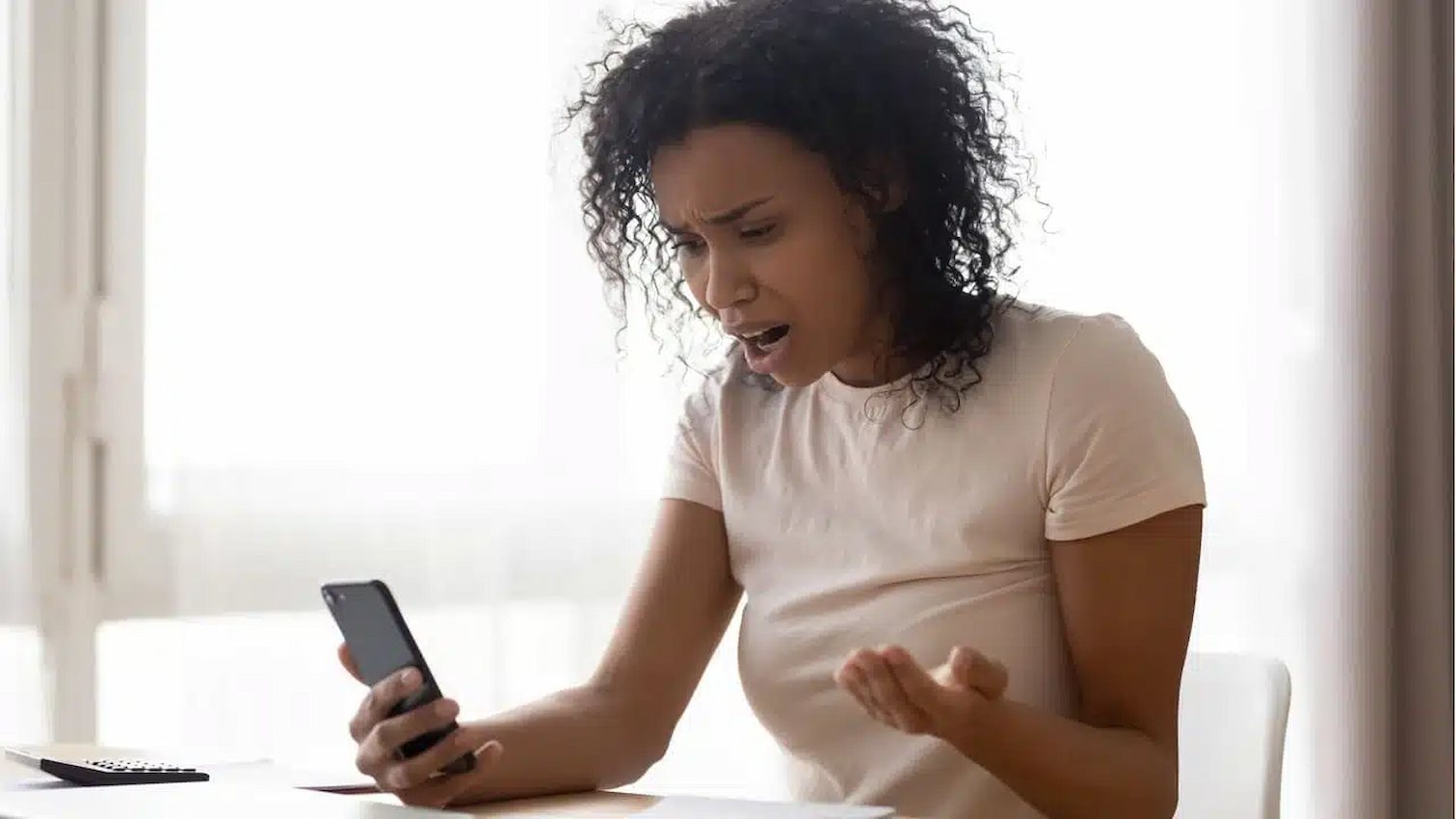 Comprar Seguidores: Como Funciona, Quais os Benefícios e os Perigos 6 Uma jovem sentada à mesa, olhando para o seu celular com uma expressão de frustração e raiva, simbolizando as possíveis decepções e problemas que podem surgir ao comprar seguidores falsos ou de baixa qualidade.