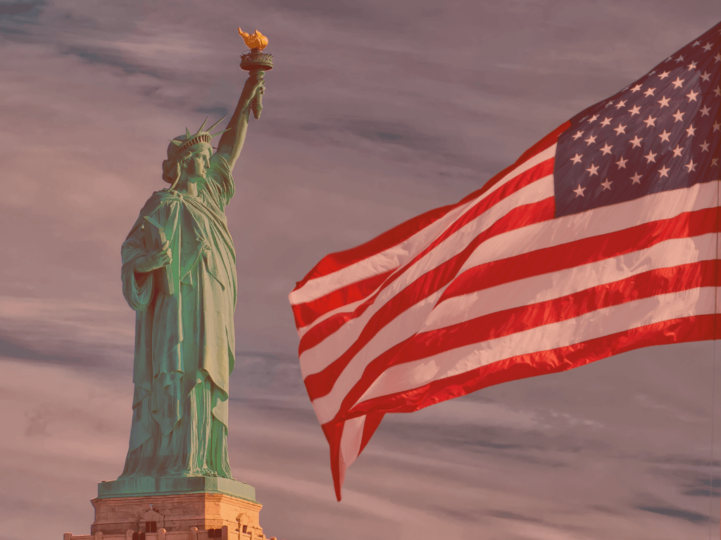 Legendas para Fotos: Frases Criativas e Inspiradoras para Instagram 2025 6 A Estátua da Liberdade em Nova York é vista em perfil, com sua tocha levantada em direção a um céu dramaticamente nublado. À direita, uma grande bandeira dos Estados Unidos tremula vigorosamente ao vento, com suas listras vermelhas e brancas e o campo azul com estrelas visíveis. A imagem evoca patriotismo, liberdade e a cultura americana, ideal para Legendas para Fotos que remetam a viagens, história ou valores nacionais.
