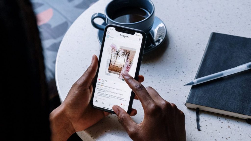 Legendas para Fotos: Frases Criativas e Inspiradoras para Instagram 2025 1 Uma pessoa segura um smartphone exibindo o feed do Instagram com uma foto de palmeiras. Ao lado, em uma mesa clara, há uma xícara de café azul e um caderno escuro com uma caneta. A imagem transmite a ideia de que a pessoa está buscando inspiração ou criando Legendas para Fotos.