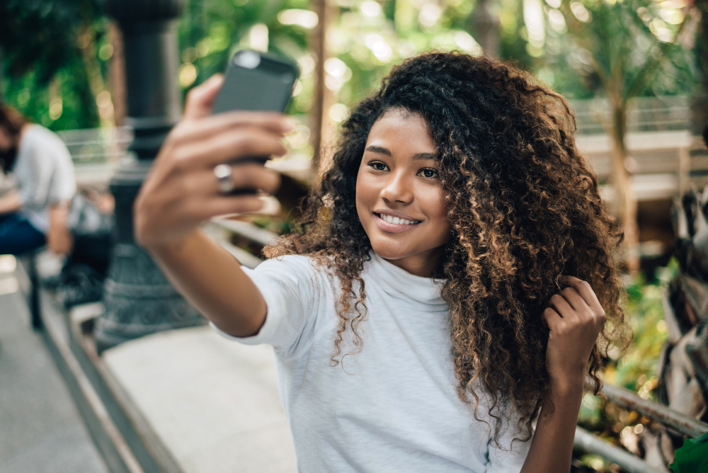 Uma mulher jovem e sorridente, com cabelos cacheados e volumosos, tira uma selfie com seu smartphone em um ambiente com folhagens verdes ao fundo. Ela veste uma camiseta branca de gola alta e parece estar em um jardim botânico ou estufa. (Legendas para Fotos)