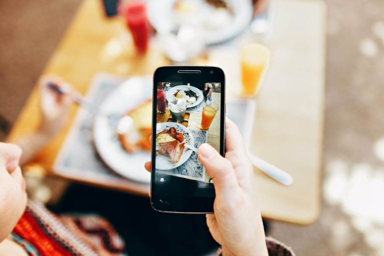 Pessoa tirando uma foto de cima de um prato de café da manhã ou brunch (com ovos, bacon, pão) com um smartphone. A tela do celular exibe a imagem que está sendo capturada. Ao fundo desfocado, há outra pessoa e uma mesa com bebidas. A cena ilustra a criação de conteúdo visual apetitoso, essencial para aumentar o alcance dos stories e engajar o público.