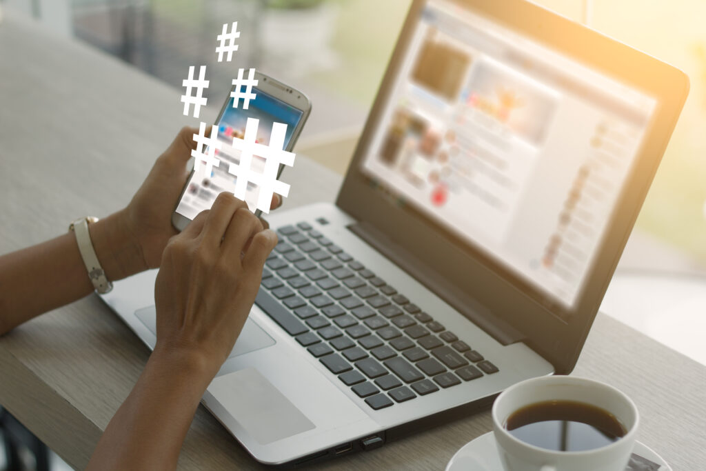 Como Ganhar Seguidores no Instagram: Estratégias Orgânicas e Dicas Infalíveis para Aumentar Seu Engajamento 3 Uma mulher está sentada em uma mesa de café, com as mãos segurando um smartphone. Na tela do telefone, ícones de hashtags (#) brancos e brilhantes flutuam, simbolizando o uso estratégico de hashtags para ganhar seguidores. Ao lado, um notebook está aberto, exibindo uma página de mídia social embaçada, e uma xícara de café está ao lado do teclado. A imagem sugere a conexão entre o uso de hashtags e o crescimento online.