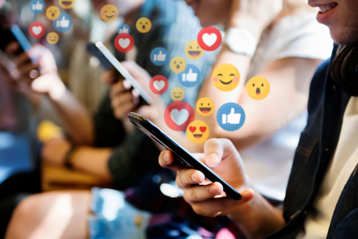 Como Ganhar Seguidores no Instagram: Estratégias Orgânicas e Dicas Infalíveis para Aumentar Seu Engajamento 4 Uma imagem de close-up mostra várias pessoas sentadas lado a lado, cada uma segurando um smartphone. Elas estão olhando para seus telefones, e ícones de curtidas, corações e emojis flutuam acima de suas mãos. A cena ilustra o engajamento e a interação online. O foco está na mão de um homem em primeiro plano, que segura o telefone. Os emojis e curtidas representam as reações e o feedback que ajudam a ganhar seguidores e aumentar o engajamento nas plataformas sociais.