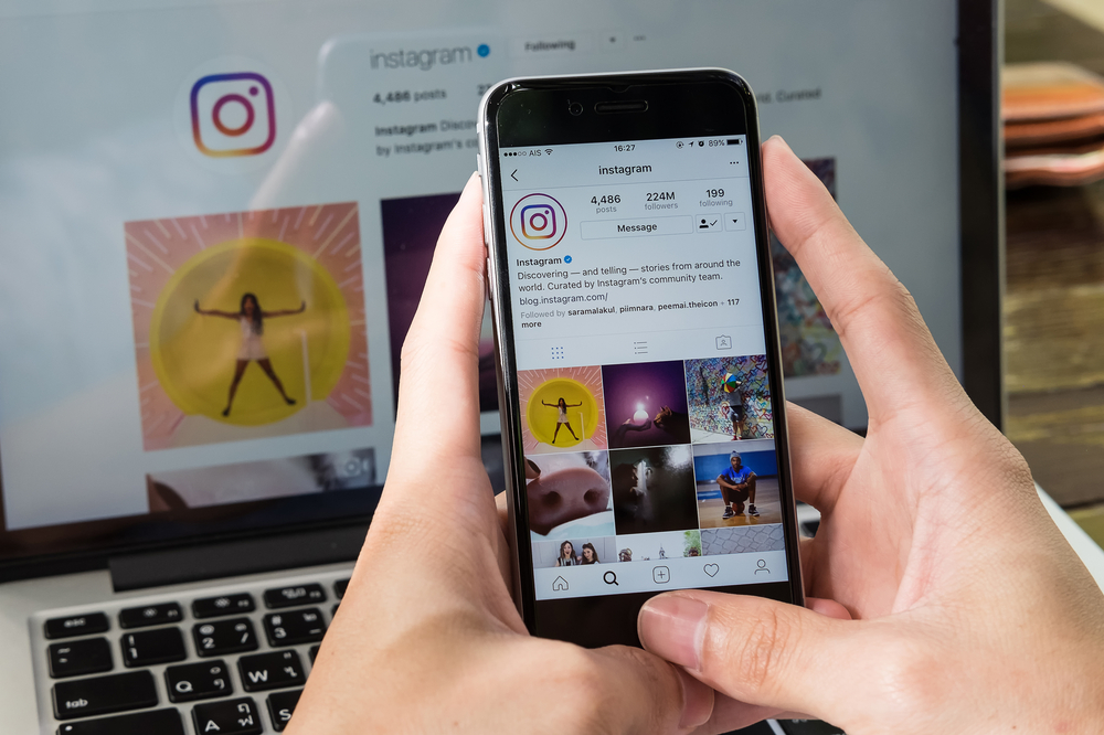 Como Ganhar Seguidores no Instagram: Estratégias Orgânicas e Dicas Infalíveis para Aumentar Seu Engajamento 5 Uma pessoa segura um smartphone na frente de um notebook, ambos exibindo a tela do Instagram. No smartphone, o perfil do Instagram com o nome de usuário "instagram" e um número de seguidores de "224M followers" está visível, e uma grade de fotos é exibida logo abaixo. A imagem demonstra como as pessoas gerenciam seus perfis e conteúdo para ganhar seguidores e engajar seu público. O foco na tela do celular e a presença do notebook sugerem um trabalho de gerenciamento de mídias sociais para alcançar o objetivo.