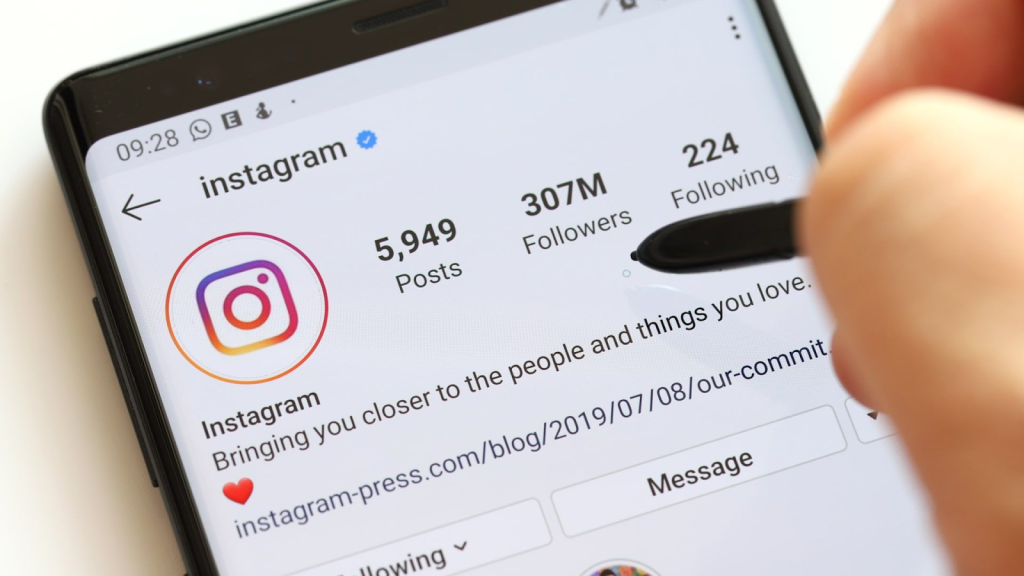 Tela de um smartphone exibindo o perfil oficial do Instagram com milhões de seguidores. Uma pessoa aponta para a tela, simbolizando o desejo de crescer na plataforma. Estratégias para ganhar visibilidade incluem criar conteúdo de qualidade e até mesmo comprar seguidores para impulsionar o engajamento.