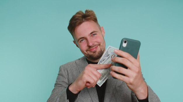 Homem de terno cinza sorridente, seguro dinheiro e um smartphone, diminuindo ganhos financeiros online e diferentes formas de ganhar dinheiro na internet.