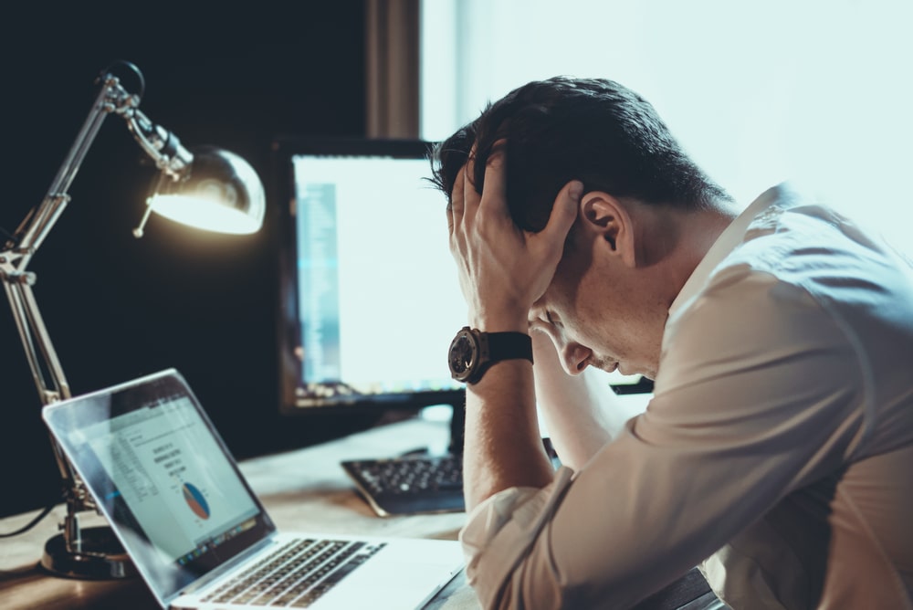 Reels para Empresas: Descubra Como Transformar Vídeos Curtos em Resultados Reais 5 Um homem, aparentemente estressado ou sobrecarregado, está sentado em uma mesa com as mãos na cabeça, em frente a um laptop e um monitor. A iluminação é baixa e uma luminária de mesa acesa adiciona foco à cena, representando os desafios na criação de Reels para empresas.