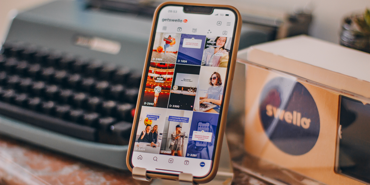 Reels para Empresas: Descubra Como Transformar Vídeos Curtos em Resultados Reais 6 Um smartphone está em um suporte, com a tela exibindo uma galeria de vídeos do Instagram Reels, com diversas miniaturas e contagem de visualizações. Ao fundo, uma máquina de escrever vintage e uma caixa com o logo "swello" simbolizam a gestão e estratégia de Reels para empresas.